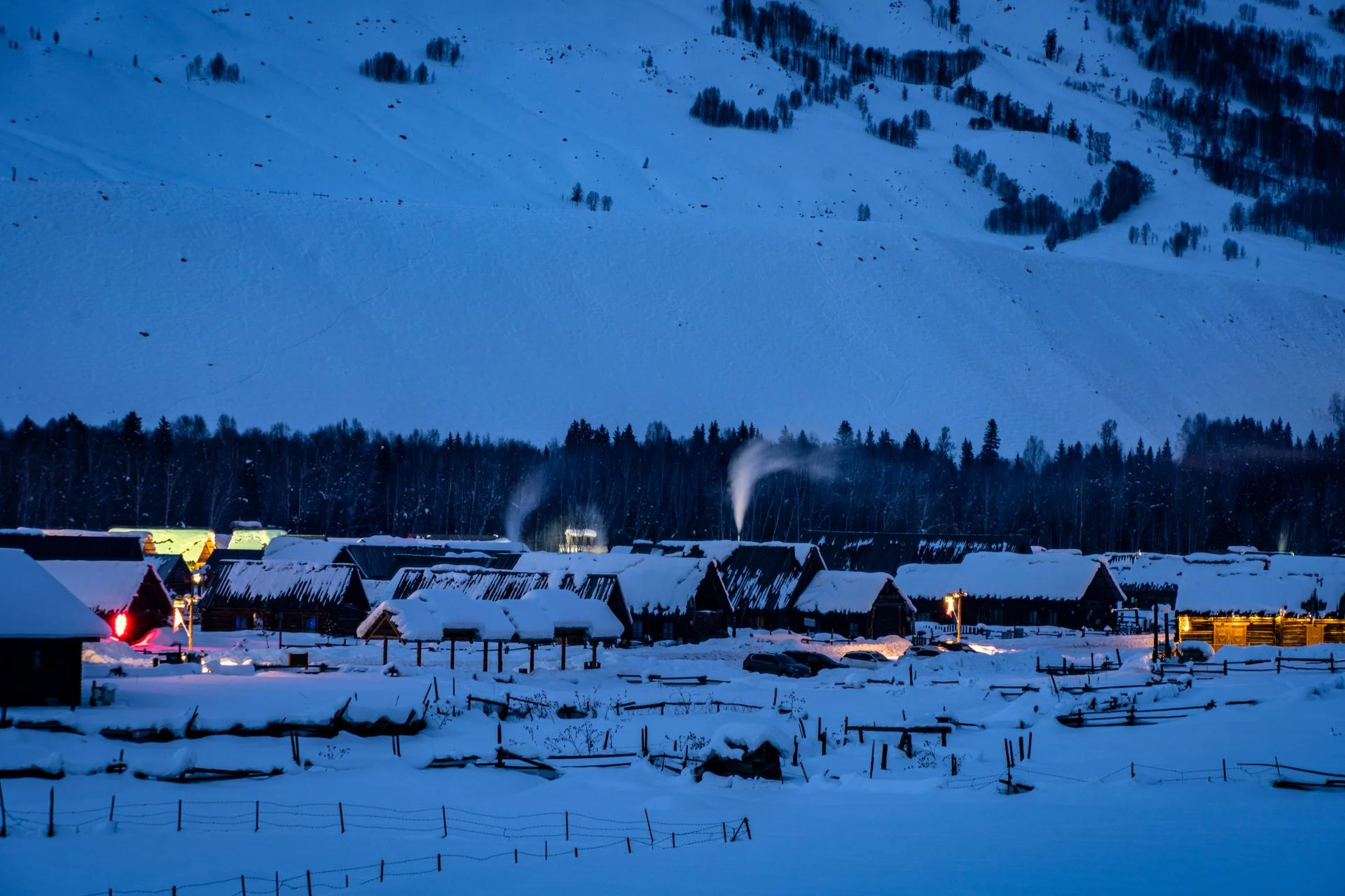 snowy village houses dusk Snow-covered Himachal village houses with warm lights at dusk below a snowy hillside
