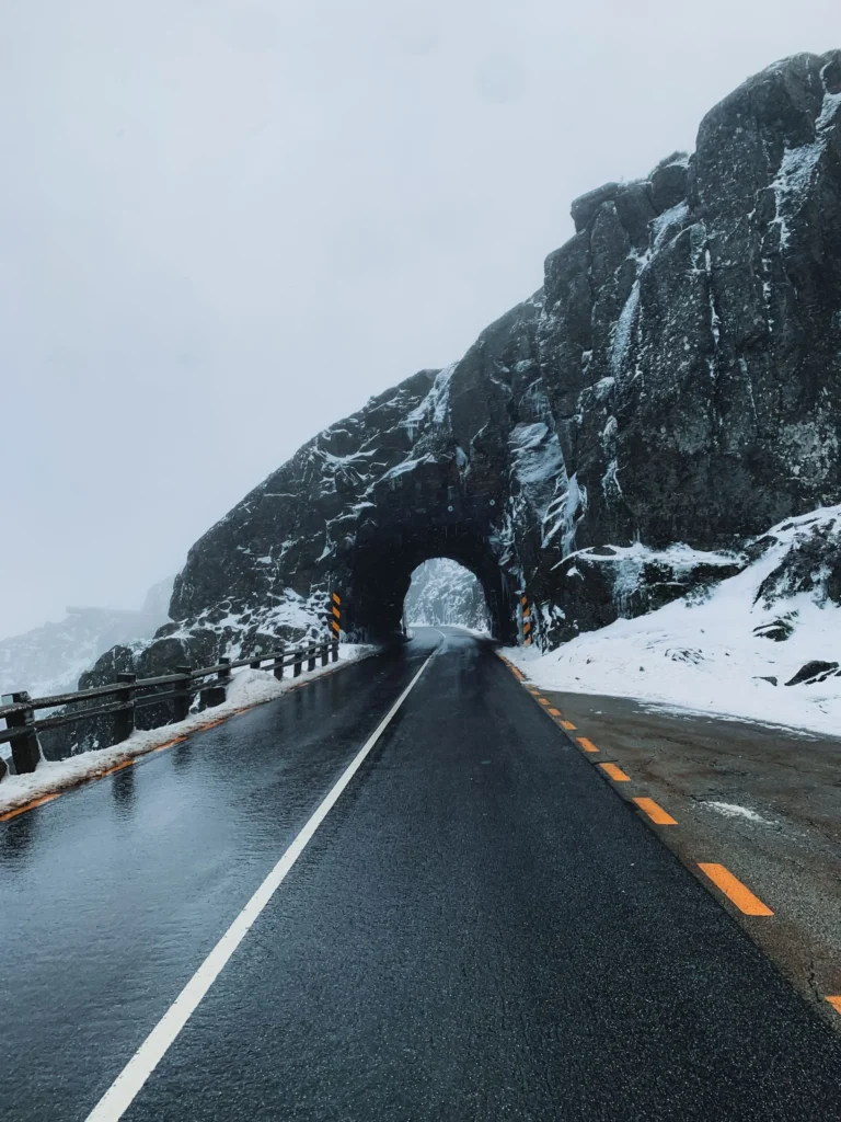 Wet mountain road leading into a short rock tunnel, with fresh snow on the cliffs and roadside